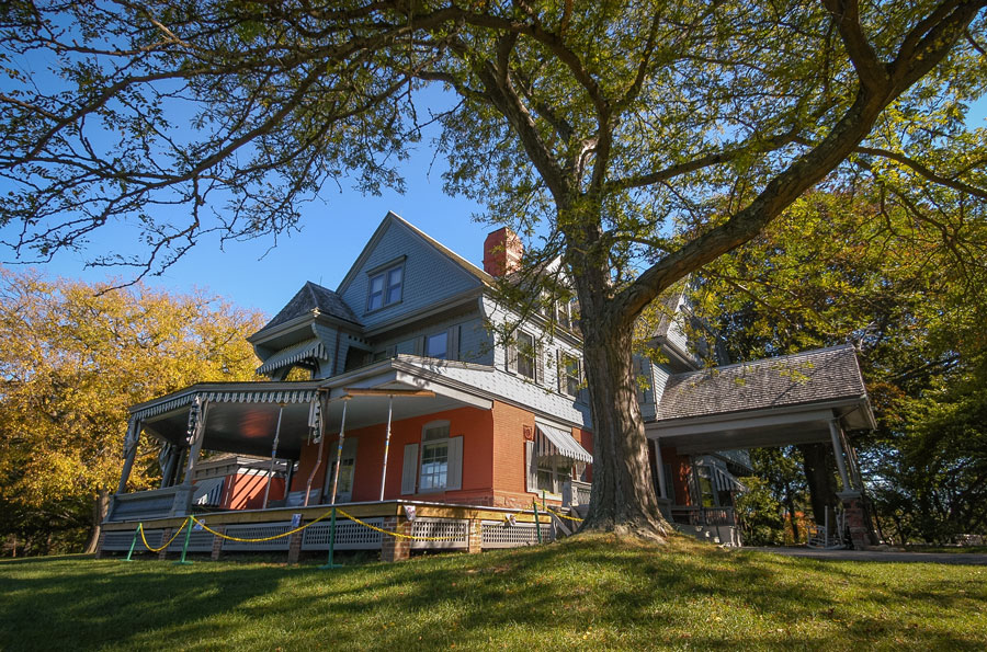 This photo shows Sagamore Hill, the historic home of President Theodore Roosevelt, framed by the branches of a large tree in the foreground. The Victorian-style residence, with its wide wraparound porch, gabled roof, and classic detailing, is set against a backdrop of vibrant green grass and early autumn foliage. The clear blue sky and warm sunlight highlight the home’s inviting architecture, making it a fitting image to accompany an article about this iconic Long Island landmark.