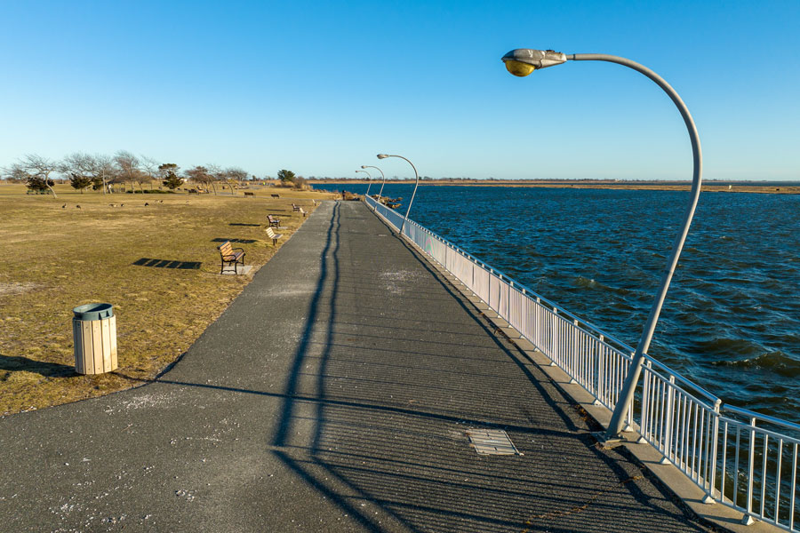 A scenic view from Wantagh Park shows a long walkway stretching along the water’s edge, framed by curved lampposts and bordered by a sturdy railing. Benches line the grassy area to the left, offering spots to relax and enjoy the peaceful waterfront. The deep blue bay glistens under the clear sky, highlighting the natural beauty and tranquility that makes Wantagh a favorite destination for outdoor recreation and waterfront leisure.