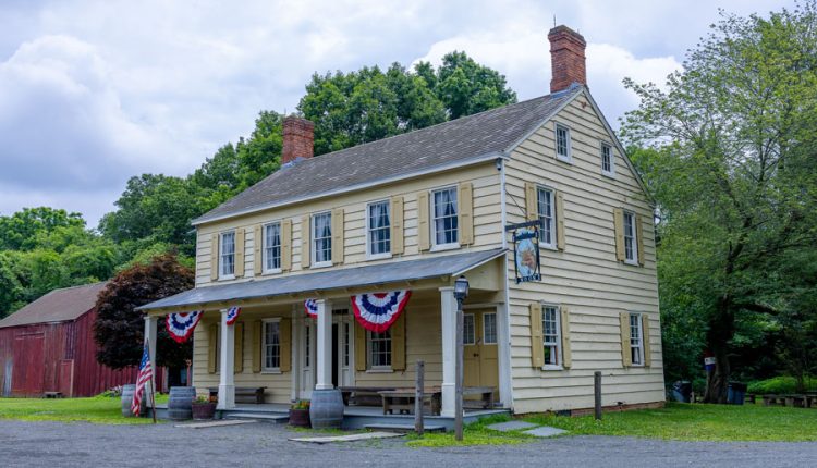 Historic colonial-style building adorned with patriotic red, white, and blue bunting, representing the type of architecture common on Long Island during the Revolutionary War era. The two-story wooden house, complete with shuttered windows, twin brick chimneys, and a front porch, evokes the character of 18th-century taverns and homesteads where early settlers lived and, in some cases, where Patriots and Loyalists gathered during the war. A weathered barn sits in the background, reinforcing the rural, agricultural setting typical of colonial Long Island.