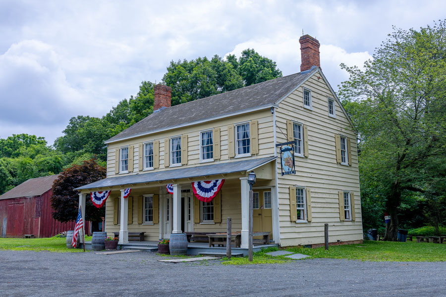 Historic colonial-style building adorned with patriotic red, white, and blue bunting, representing the type of architecture common on Long Island during the Revolutionary War era. The two-story wooden house, complete with shuttered windows, twin brick chimneys, and a front porch, evokes the character of 18th-century taverns and homesteads where early settlers lived and, in some cases, where Patriots and Loyalists gathered during the war. A weathered barn sits in the background, reinforcing the rural, agricultural setting typical of colonial Long Island.