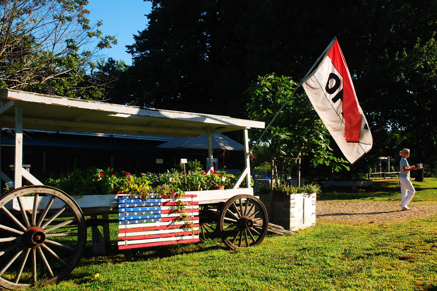 A local farmers market in Amagansett, Long Island, New York, photographed on July 26. A rustic wagon decorated with an American flag planter and an Open banner welcomes visitors to shop for fresh, organic produce and locally grown goods, reflecting the Hamptons’ farm-to-table culture.