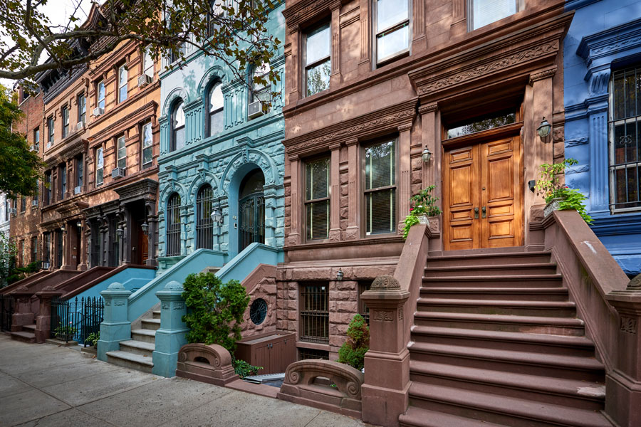 A picturesque row of historic brownstone townhouses, showcasing the classic stoops, ornate facades, and architectural charm that define many of Manhattan’s iconic neighborhoods. The colorful mix of stone and painted exteriors highlights the city’s blend of tradition and individuality, while tree-lined sidewalks add to the residential character of this urban streetscape.