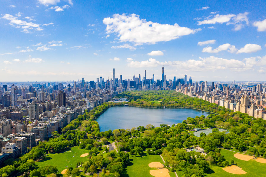 A stunning aerial view of Central Park in New York City, showcasing the Jacqueline Kennedy Onassis Reservoir, scenic green spaces, and baseball fields, with the iconic Manhattan skyline rising in the background. A must-visit destination blending nature and city life.