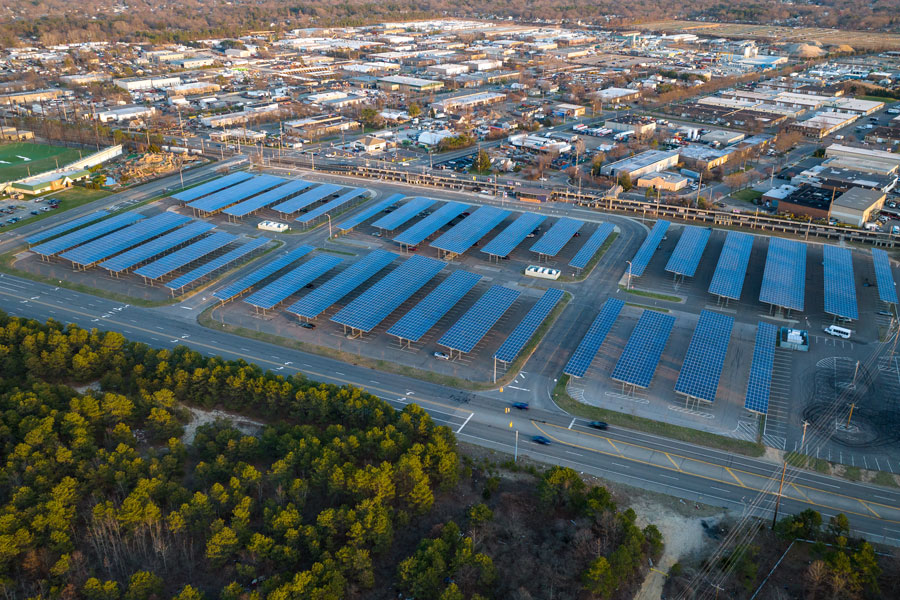 The Deer Park Long Island Railroad station parking lot, showcasing an array of car shelters with solar panels installed on their rooftops. Surrounding the parking lot, a mix of suburban commercial buildings and residential neighborhoods stretches into the distance, while a wooded greenbelt frames the lower portion of the image.