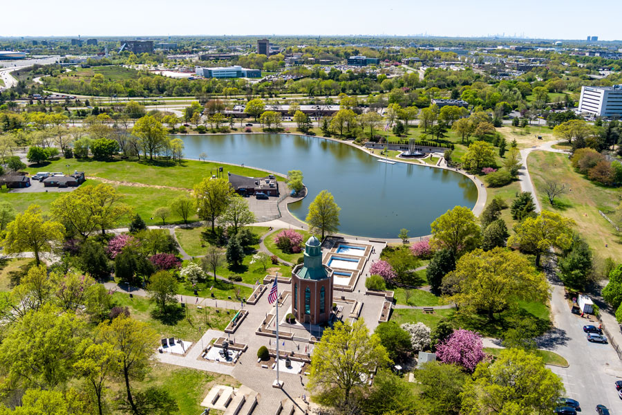Aerial view of Eisenhower Park in East Meadow, Nassau County, New York. The photo highlights the park’s central lake surrounded by lush greenery, spring blossoms, and walking paths. In the foreground is the Veterans Memorial Tower with the American flag, a key landmark within the park. Beyond the park, the suburban landscape of Long Island stretches toward the horizon. 