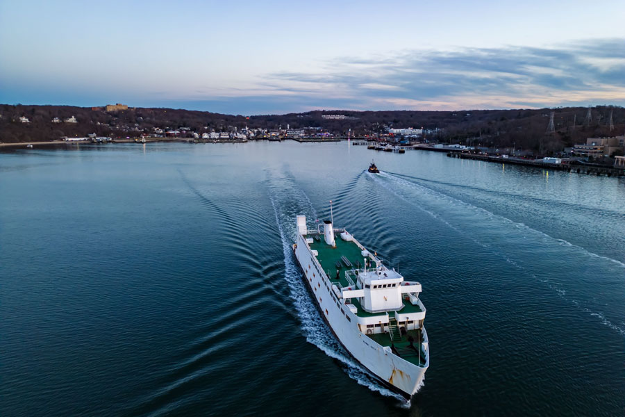 The Bridgeport & Port Jefferson Ferry departing Connecticut en route to Long Island, New York,. The vessel is seen moving across the calm waters of Port Jefferson Harbor, leaving a wake behind it as it heads out to Long Island Sound. On the shoreline, the village of Port Jefferson is visible, with its clustered buildings, hills, and waterfront activity. The ferry has been a vital transportation link between Port Jefferson, NY, and Bridgeport, CT for over a century, carrying both passengers and vehicles across the Sound.