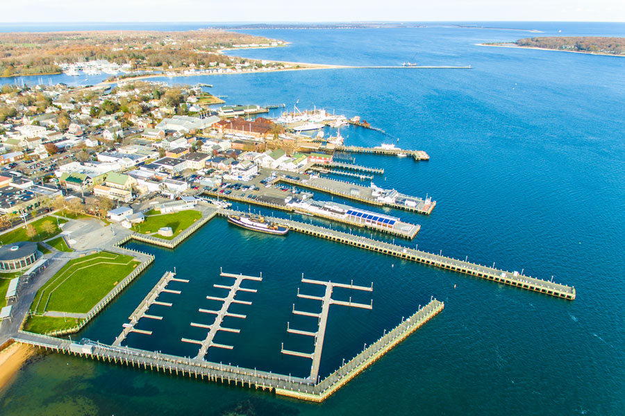 An aerial view of Gardiners Bay and its harbor area, located on the eastern end of Long Island, New York. The photo highlights the waterfront, docks, and marina infrastructure, with a town clustered around the shoreline and extending inland. Gardiners Bay is known for its scenic beauty, maritime activity, and its role as a gateway to the East End of Long Island, including popular destinations like Greenport, Shelter Island, and Montauk.