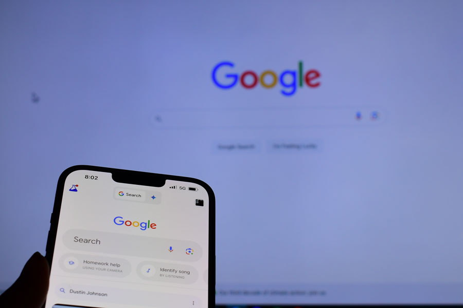A smartphone displays the Google search app in front of the Google homepage on a computer screen. The tech giant avoided a breakup in a major antitrust ruling but was ordered to share data with rivals.