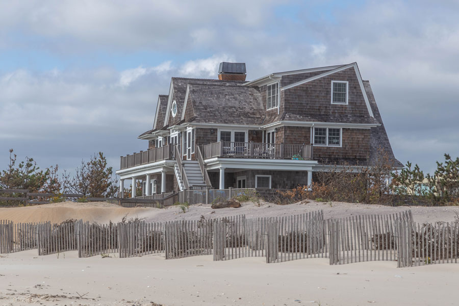 A classic shingle-style beach house sits behind the dunes in the Hamptons, Long Island, New York. With weathered cedar siding, wraparound porches, and views of the Atlantic Ocean, this architectural style reflects the timeless coastal charm of the Hamptons