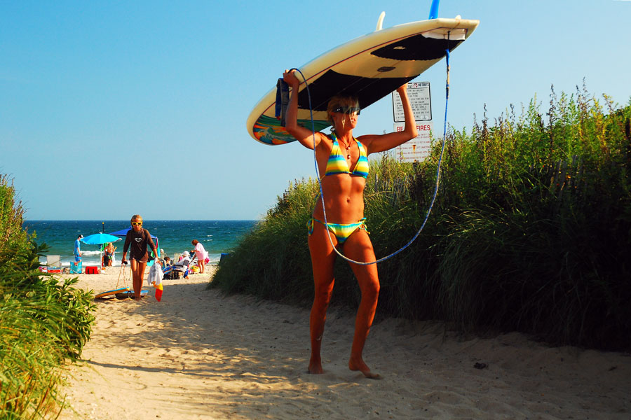 A surfer carries her board from the beach on a summer day in the Hamptons, Long Island, New York. The region is known for its pristine Atlantic beaches, with Ditch Plains in Montauk recognized as one of the premier surfing destinations on the East Coast.
