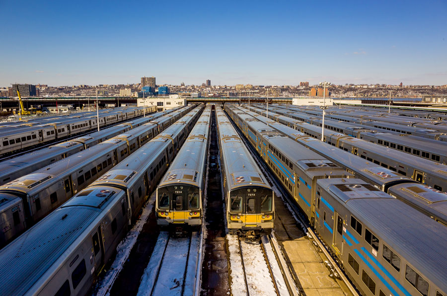 This photo shows a Long Island Rail Road (LIRR) train yard filled with rows of commuter trains parked side by side. The silver train cars with their yellow-and-black fronts are the M7 and M9 electric multiple units used daily by thousands of riders. Train yards like this are essential for storing, maintaining, and preparing trains between runs. The urban skyline in the background highlights the LIRR’s vital role in connecting Long Island with New York City.