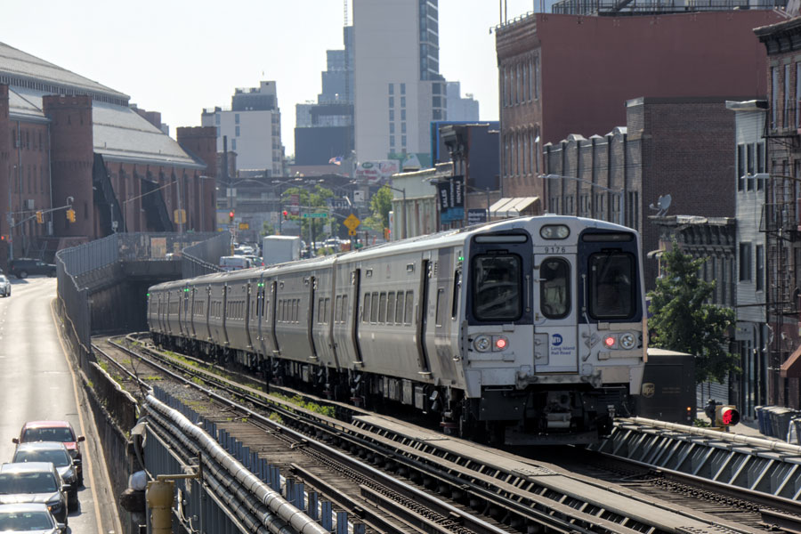 A Long Island Rail Road (LIRR) commuter train traveling past the elevated Nostrand Avenue station in Brooklyn, New York, with busy Atlantic Avenue running parallel to the tracks. The photo, taken on June 4, 2025, captures the urban landscape with its mix of historic and modern architecture, dense residential and commercial buildings, and active rail and street traffic. 