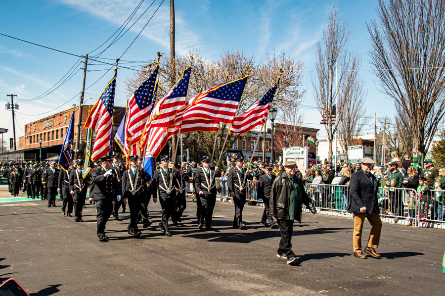 Lindenhurst, New York, USA, March 30, 2024 - Uniformed Marchers Carrying American Flags Walk Proudly Down A Sunny Street During A Parade, With A Lively Crowd Of Spectators Lined Up On The Sides