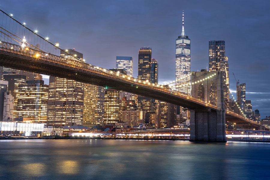 This stunning nighttime view captures the Brooklyn Bridge stretching across the East River, its lights forming a glowing pathway that leads toward Manhattan’s glittering skyline. The reflective waters below add depth to the scene, while the soaring One World Trade Center stands tall among the surrounding skyscrapers, symbolizing resilience and modern New York. The blend of historic architecture and contemporary towers showcases the city’s dynamic character, making this one of the most recognizable and photographed vistas in the world.