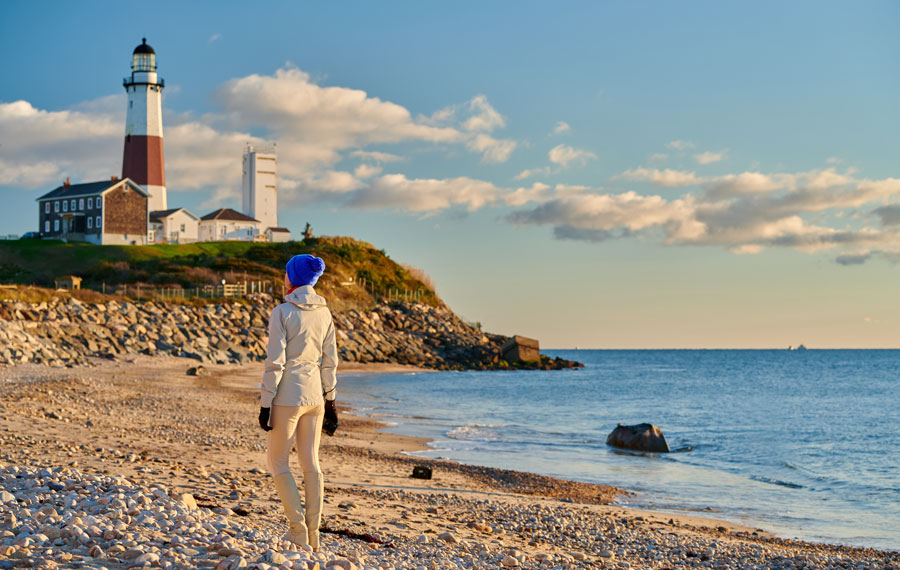 A visitor enjoys a peaceful stroll along the rocky beach near Montauk Point Lighthouse, the oldest lighthouse in New York State. Situated at the easternmost tip of Long Island, this historic landmark has guided mariners since 1796 and stands as a symbol of maritime heritage against the backdrop of the Atlantic Ocean.