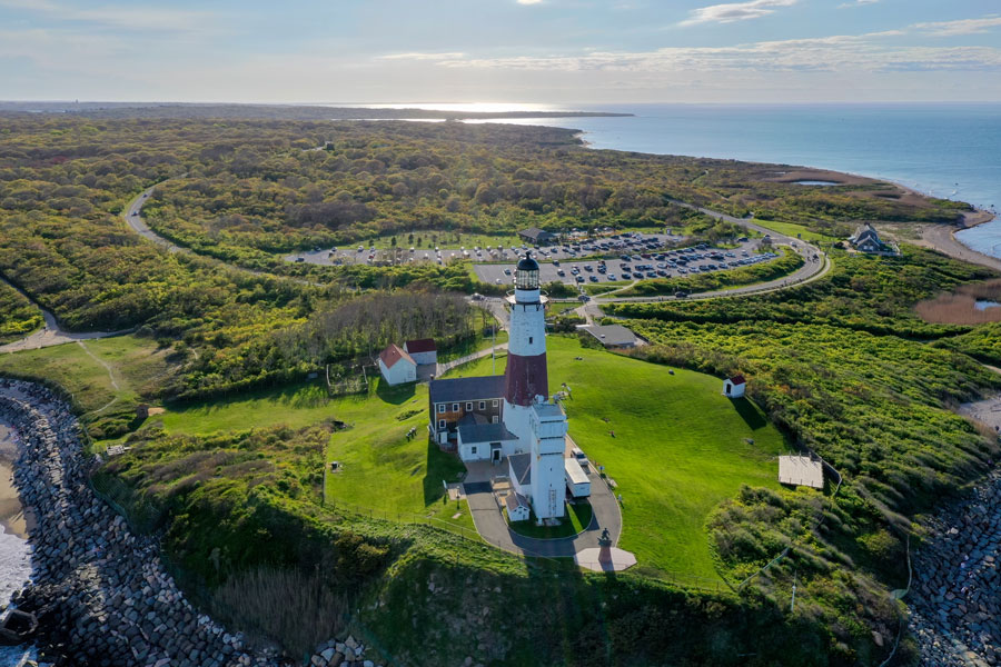 A breathtaking aerial view of the historic Montauk Point Lighthouse, perched atop the easternmost tip of Long Island, New York, surrounded by lush greenery and overlooking the vast Atlantic Ocean.