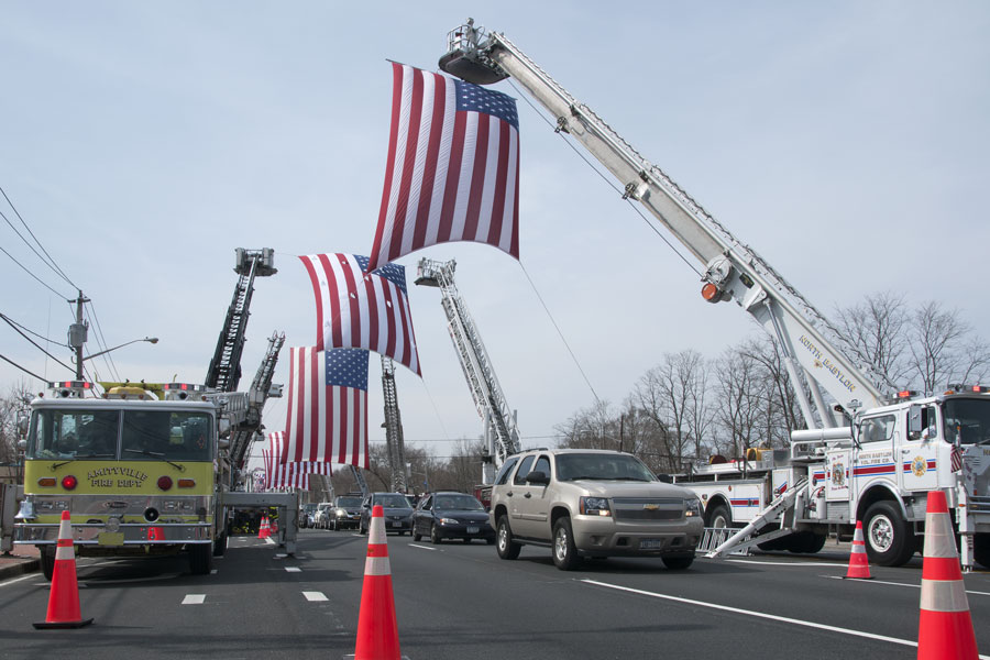 Fire departments line Middle Country Road in Selden, NY, with ladder trucks displaying large American flags on March 18, 2011, in preparation for the funeral procession of NYPD Officer Geoffrey Breitkopf, who tragically died in a friendly fire incident. The tribute reflects the strong sense of community and solidarity in Selden during times of loss.