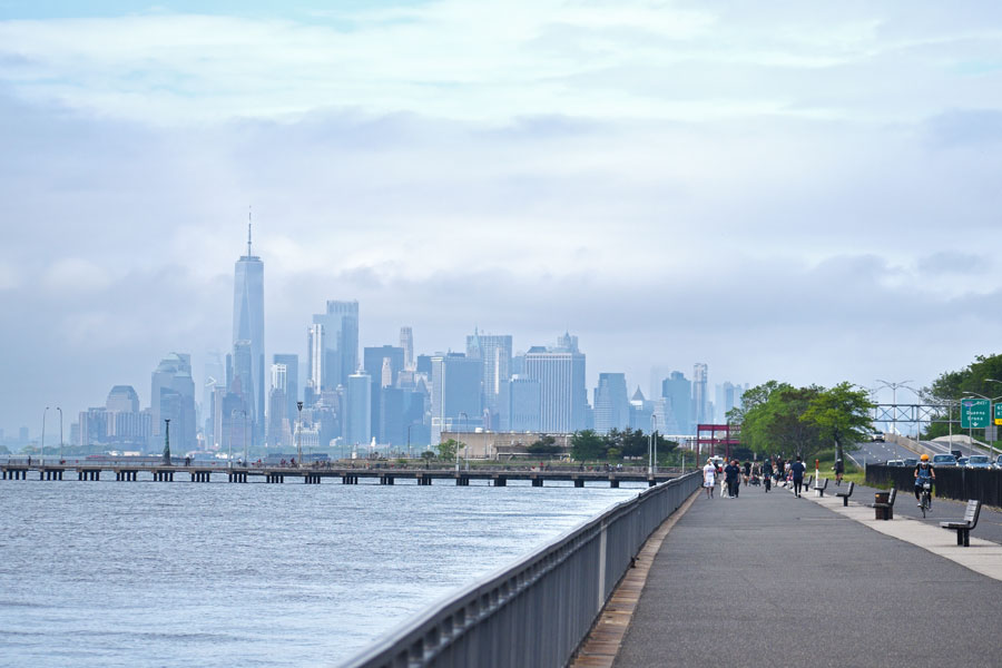 The image shows the Belt Parkway Promenade in Bay Ridge, Brooklyn, with people walking and biking along the waterfront. In the distance, the Manhattan skyline is visible through the clouds, including One World Trade Center. This promenade does connect neighborhoods within Brooklyn, but to get to other parts of Long Island, you’d need to continue eastward into Queens and beyond.