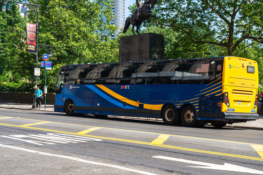 A New York City MTA bus stopped near Central Park. The bus features the system’s bold blue-and-yellow livery accented by the I ♥ NY logo, symbolizing the pride of the city’s transit network. A banner promoting a Broadway show and a nearby bus stop sign add to the urban setting. Reflections of Manhattan’s tall buildings gleam across the bus windows, reminding us that this service links neighborhoods across all five boroughs.