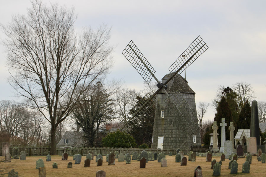The historic North End Cemetery in East Hampton, NY, with the iconic Old Hook Mill, a well-preserved 19th-century windmill, in the background. The cemetery, dating back to colonial times, contains the graves of early settlers and veterans, reflecting the rich heritage and enduring legacy of the East Hampton community. The scene highlights the town’s blend of agricultural history and memorial tradition, preserved in this unique setting.