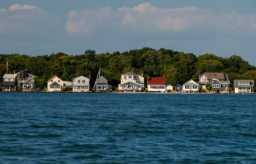 This image shows a panoramic view of Greenport, Long Island, New York, taken from the water while sailing. A row of charming waterfront homes lines the shore, each with its own unique architecture and direct access to the bay. Behind the houses, a backdrop of lush, green trees creates a peaceful coastal setting. Greenport is a historic maritime village on the North Fork, known for its scenic harbor, boating culture, and vibrant waterfront community.