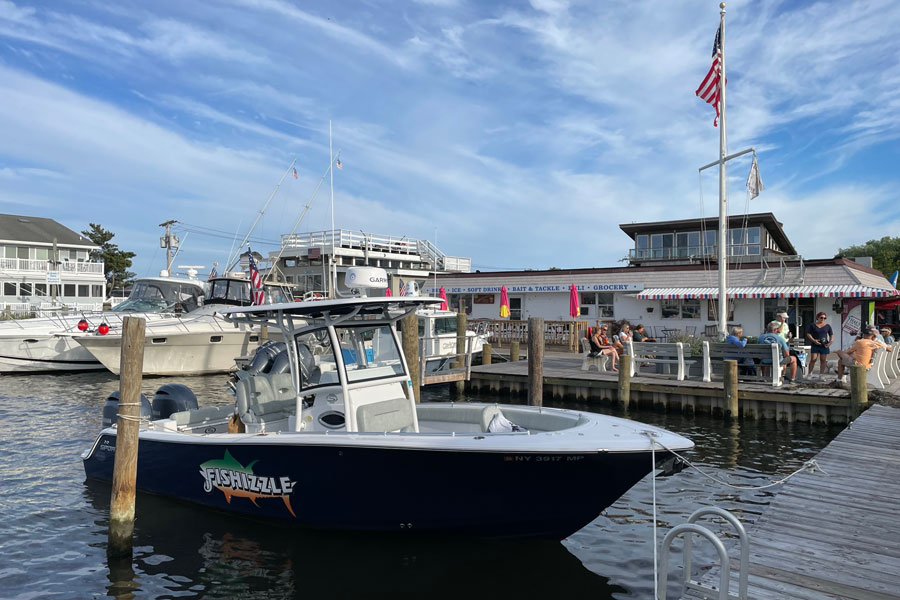 A small fishing boat moored in the harbor at Kismet, Fire Island, New York, on September 16, 2023. The lively waterfront scene shows visitors relaxing at a café by the docks, reflecting the laid-back beach-town atmosphere that makes Kismet a popular summer destination.