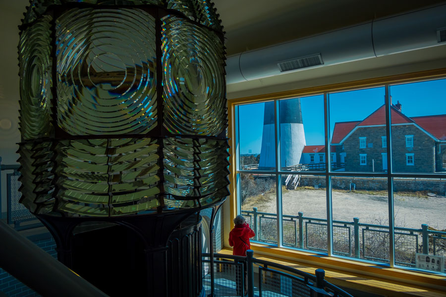 This photograph captures the magnificent Fresnel lens exhibited inside the Montauk Point Lighthouse Museum. Taken on April 14, 2018, the image offers an interior perspective of the immense, intricately designed lens, an engineering marvel that played a vital role in guiding ships safely past Long Island's treacherous shores. Beyond the lens and through the museum window, you can see the historic lighthouse tower and the adjoining keeper’s buildings.