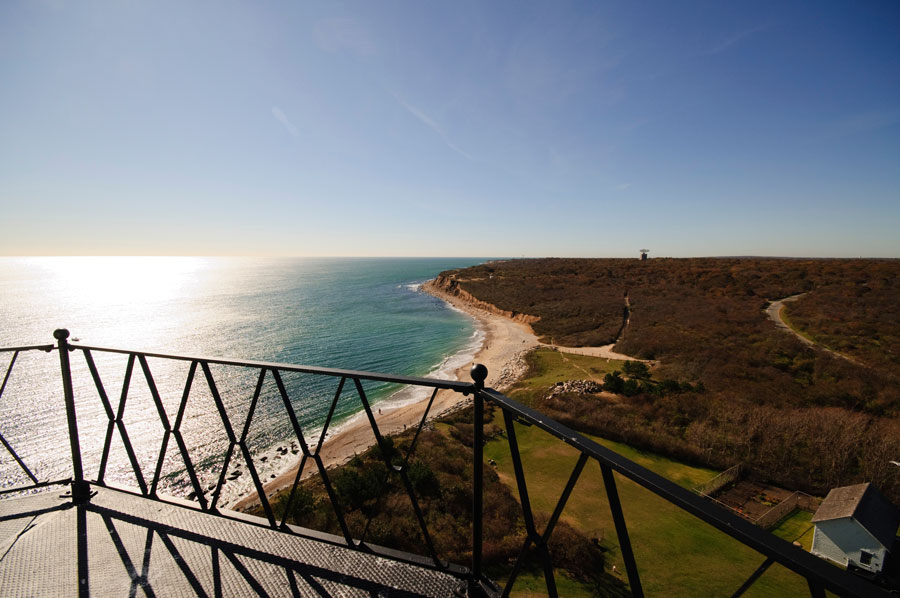 The stunning view from Montauk Point Lighthouse, looking out over the Atlantic Ocean and the rugged coastline of the eastern tip of Long Island, New York. The vantage point from the lighthouse offers panoramic vistas of the beach below, the glimmering water, and the natural landscape stretching toward the horizon, making it a unique and iconic sight associated with Montauk.