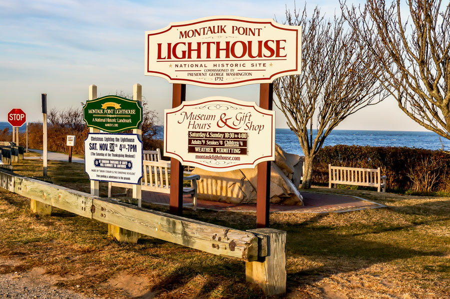A welcoming sign at the entrance to Montauk Point Lighthouse, a national historic site commissioned by President George Washington in 1792. The sign details museum and gift shop hours, inviting visitors to explore the landmark overlooking the Atlantic Ocean.