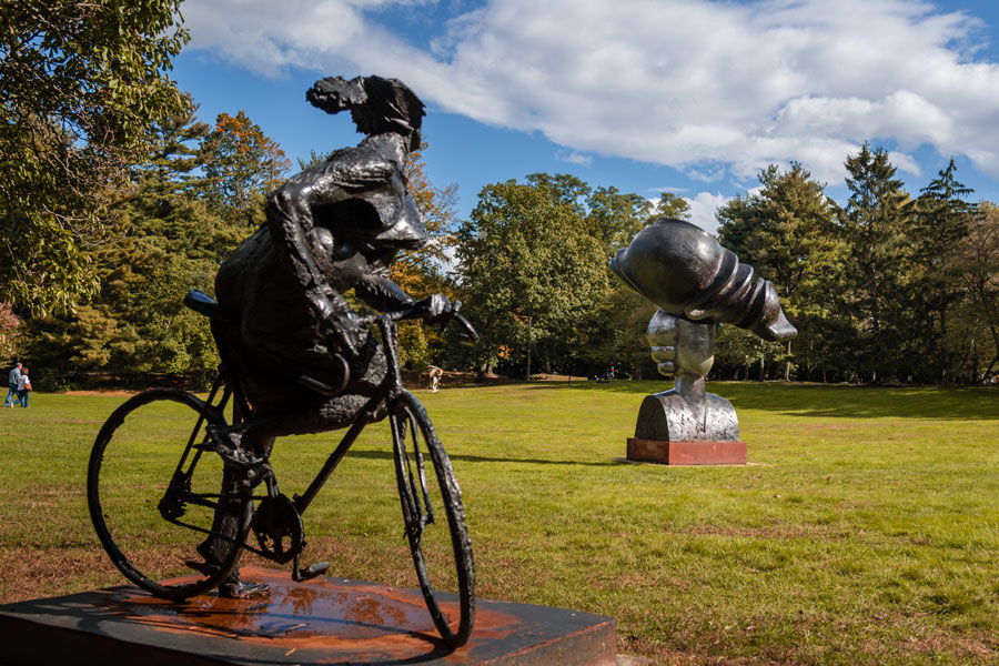two prominent outdoor sculptures in a park setting in Roslyn, New York. In the foreground, there is a sculpture of a woman riding a bicycle, rendered in an expressive, textured style, and in the background, another sculpture depicts a woman's head wearing an oversized hat, held by a sculpted hand, with highly polished metallic surfaces.
