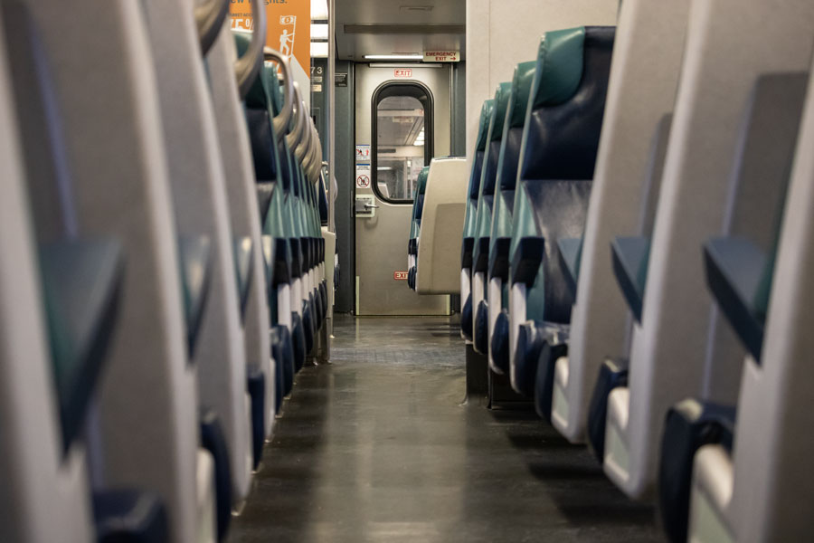 This interior seating arrangement of a Long Island Rail Road (LIRR) commuter rail train. The photograph is taken from aisle-level, looking toward the exit door at the end of the rail car, with rows of blue and grey seats on either side. Ample legroom and straightforward access to exits accommodate high passenger turnover typical of commuter rail services. 