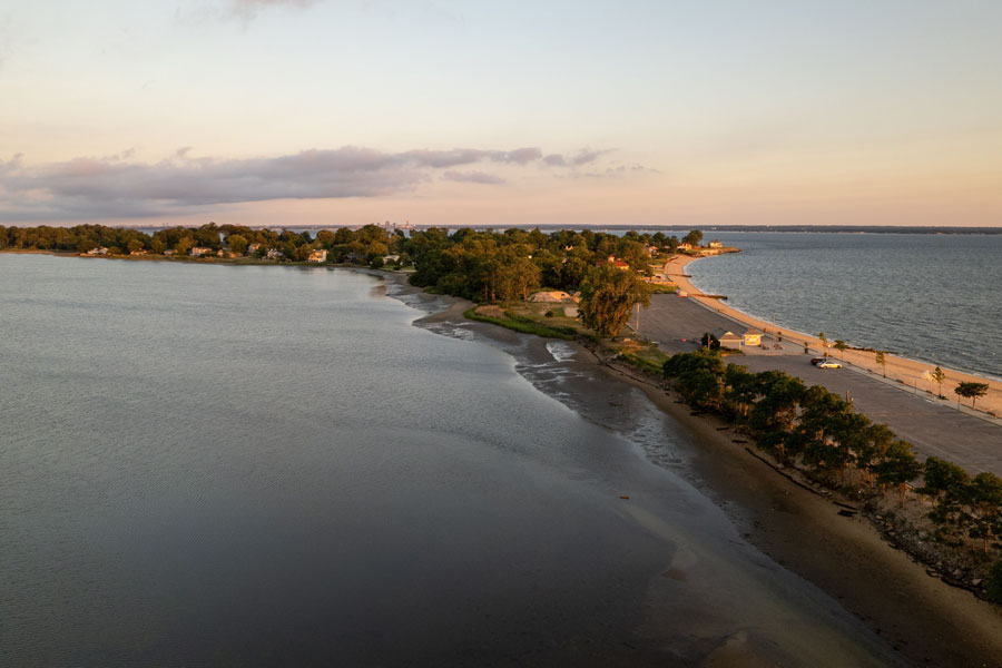Aerial view of a golden sunrise along the Gold Coast of Long Island, New York. The early morning light casts a warm glow over the shoreline and calm waters, illuminating the quiet beach and tree-lined coast, showcasing the area’s serene natural beauty.