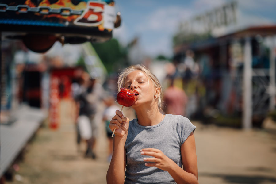 A young woman enjoying a red candy apple at an outdoor fair, with food stands and festival booths in the background on a sunny day at the Town of Islip Apple Festival in Sayville, New York.