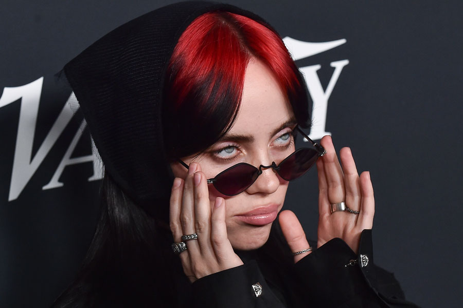 Billie Eilish on the red carpet wearing dark sunglasses and a black outfit with red and black hair at Variety’s Power of Women event in Hollywood.