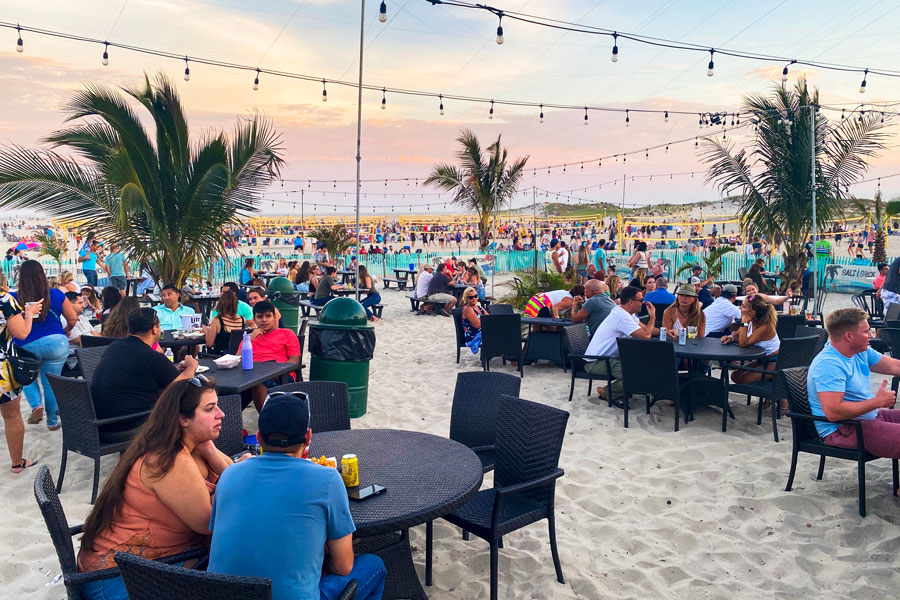 People dining and socializing at tables on the sand at Cedar Beach in Babylon, New York, with palm trees, string lights, and volleyball games taking place in the background on a warm summer evening.