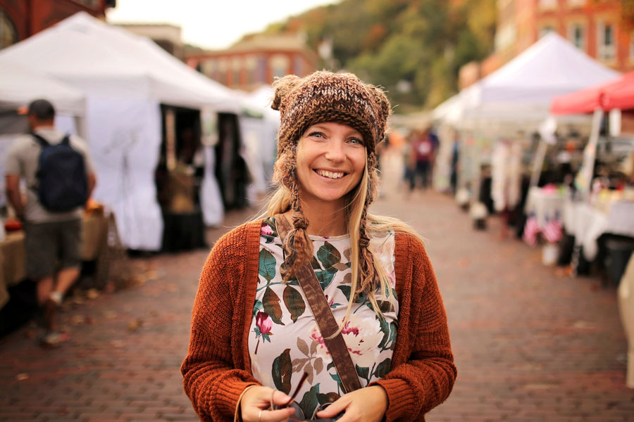 A smiling woman in a cozy autumn outfit visits a Long Island street fair lined with vendor tents during the fall festival season.