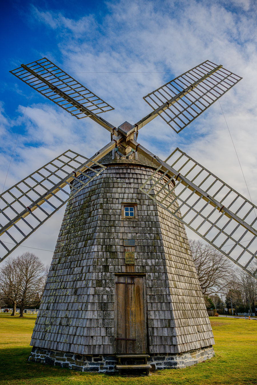 Front view of the James Corwith Grist Windmill in Water Mill, New York, showing its tall shingled tower, four wooden lattice sails, and stone base beneath a bright blue sky.