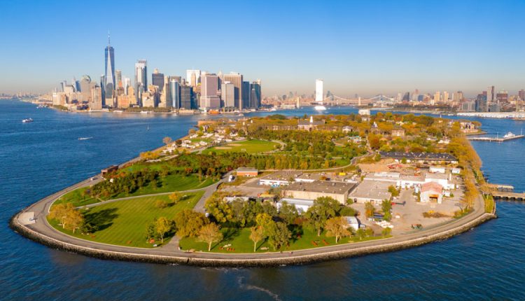 Aerial photograph of Governors Island in New York Harbor showing its green parkland surrounded by water, with the skyscrapers of Lower Manhattan and the Brooklyn and Manhattan Bridges visible in the distance under a clear blue sky.