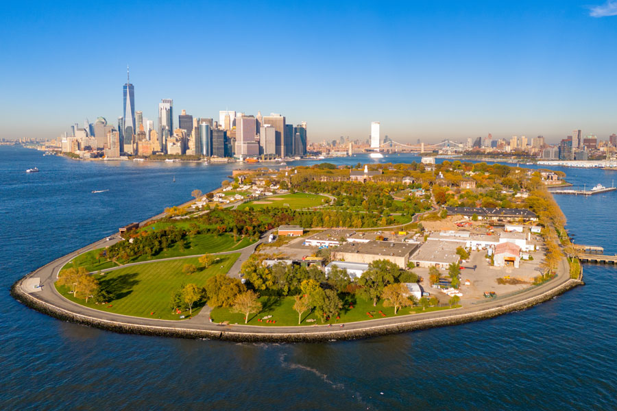 Aerial photograph of Governors Island in New York Harbor showing its green parkland surrounded by water, with the skyscrapers of Lower Manhattan and the Brooklyn and Manhattan Bridges visible in the distance under a clear blue sky.