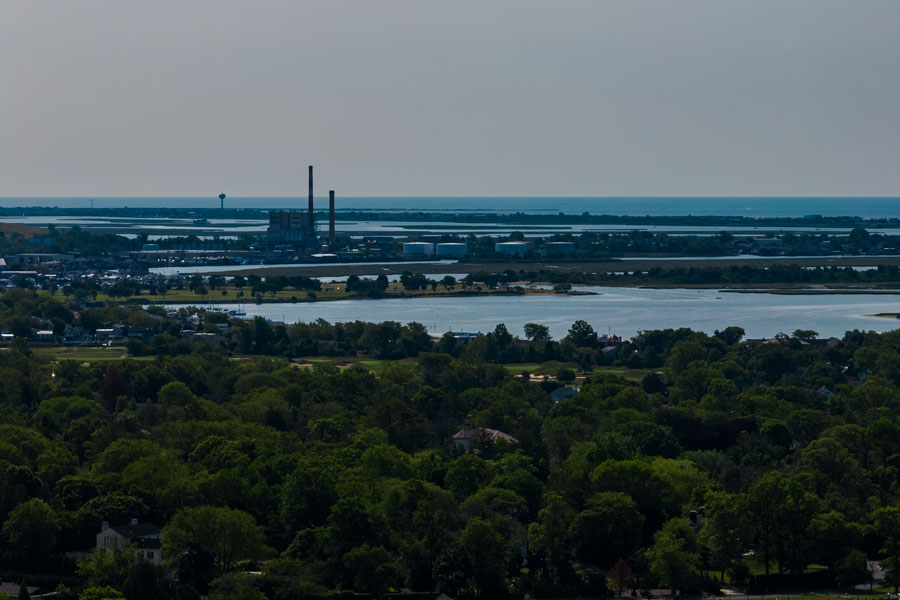 A high-altitude aerial view overlooking Hewlett, New York, captures the community lush suburban canopy with Long Beach visible in the hazy distance. The image illustrates Hewlett’s unique geography within Nassau County Five Towns, where quiet tree-lined neighborhoods meet the open waterways leading toward the Atlantic Ocean. The shot also highlights the area’s proximity to both serene residential landscapes and the broader South Shore coastal environment.
