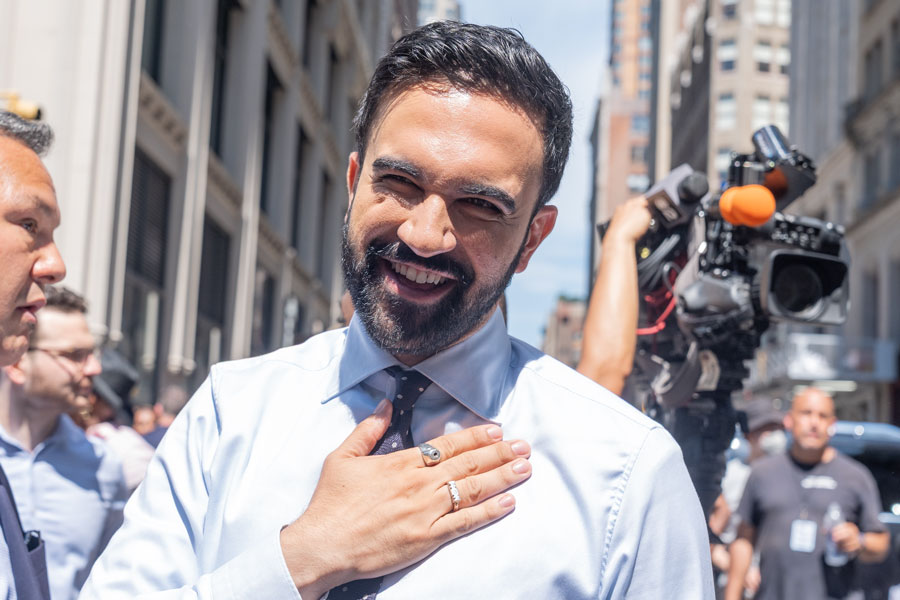 Zohran Mamdani smiling and placing his hand on his chest while greeting supporters outside in New York City after the first 2025 mayoral debate, with news cameras and downtown buildings in the background.