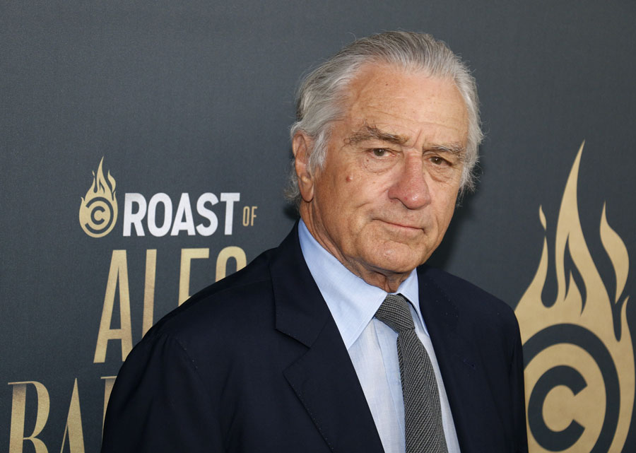 Robert De Niro in a navy suit at a red-carpet event, standing before a backdrop reading “Roast of Alec Baldwin.