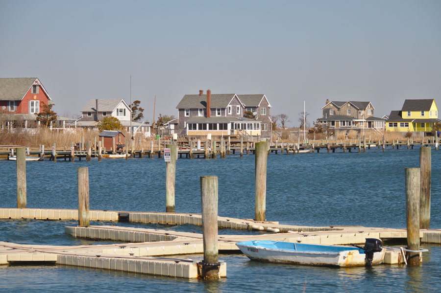 Waterfront homes and private docks on Oak Island, Suffolk County, New York, with a small motorboat tied to floating piers under a clear blue sky on a calm early spring day.