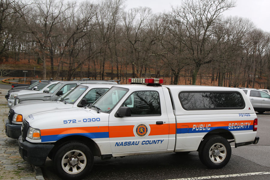 Nassau County Public Security vehicle parked at Plainview-Old Bethpage Community Park with trees and parked cars in the background on an overcast day.