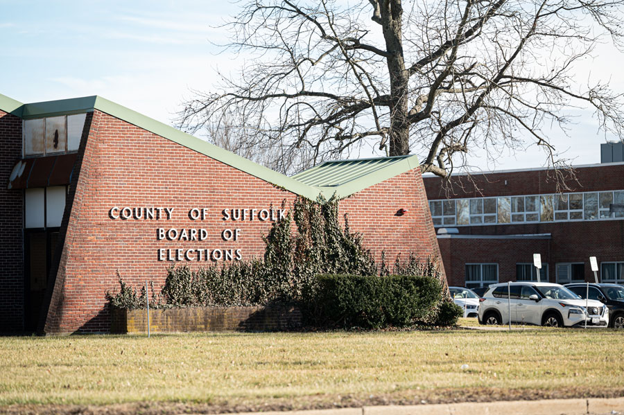 The Suffolk County Board of Elections building in Yaphank, New York, features a distinctive red-brick exterior with a sloped green roofline. The facility serves as the central hub for election administration and voter services in Suffolk County. Several vehicles are parked nearby, and leafless trees frame the scene on a clear day.