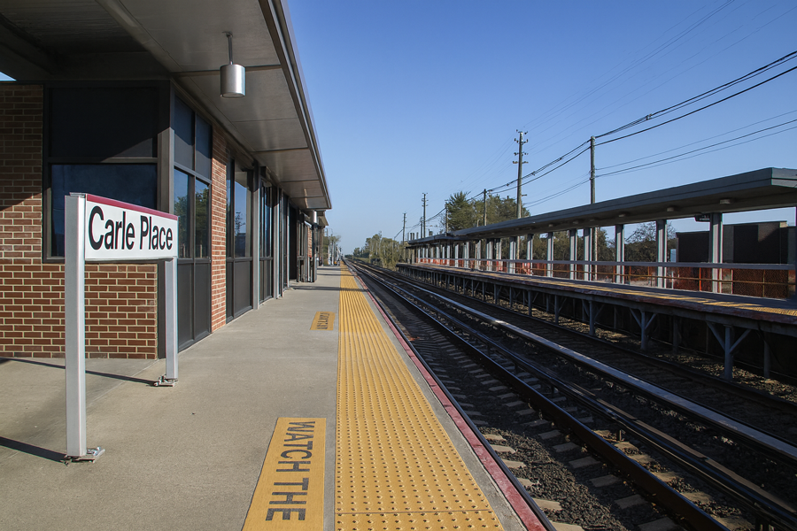Carle Place LIRR station platform on a sunny day in Nassau County, NY, showing tracks, signage, and station canopy surrounded by trees.