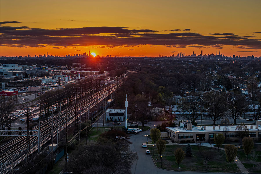 A golden sunrise rises over Valley Stream, Long Island, with the Green Acres Shopping Mall and its neighboring commercial district visible along Sunrise Highway. The early morning light captures the balance between suburban life and proximity to New York City, whose skyline stretches dramatically across the horizon. This vantage highlights Green Acres Mall’s position as a major retail hub connecting Nassau County and the city beyond.