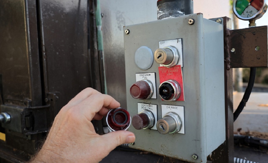 Close-up view of a person’s hand inspecting the control buttons on an industrial trash compactor, showing safety switches and indicator lights.