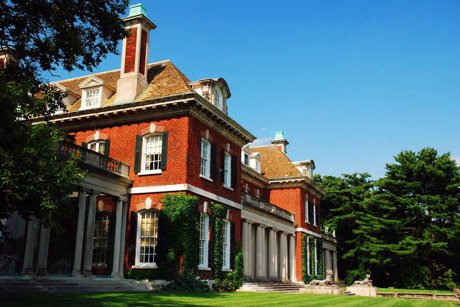 Historic brick mansion in Old Westbury, New York, featuring ivy-clad walls, white columns, and landscaped lawns under a bright blue summer sky.