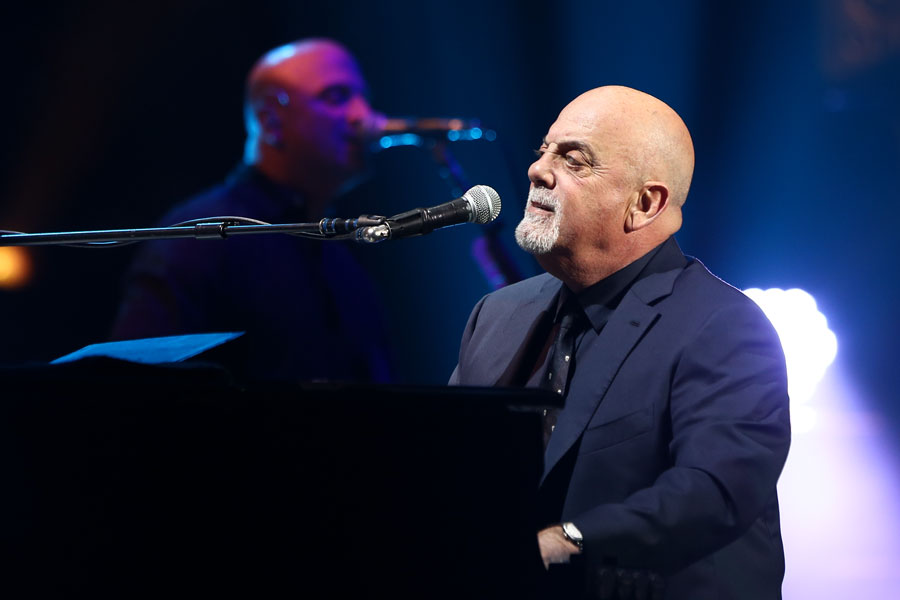Billy Joel performing at Madison Square Garden in 2016, singing at the piano under stage lights.